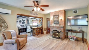 A living room with a fireplace, a ceiling fan, a TV, and a kitchen in the background. - Cozy Mars Hill Farmhouse w/ Scenic Views (Mars Hill)