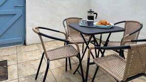 Un petit espace extérieur aménagé avec des chaises et une table en osier, une machine à café et des croissants.