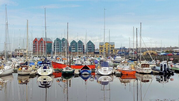 Un port de plaisance où de nombreux bateaux sont amarrés, des bâtiments aux couleurs vives en arrière-plan et une eau calme qui reflète les bateaux.