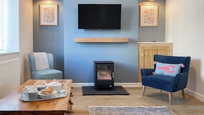 Un salon avec un téléviseur fixé au mur, une cheminée, un canapé, un fauteuil et une table basse sur laquelle se trouve un plateau de pâtisseries.