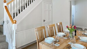 Un coin repas avec une table en bois dressée pour quatre personnes, des chaises blanches et un vase de tulipes. Une porte blanche et un escalier sont visibles à l’arrière-plan.