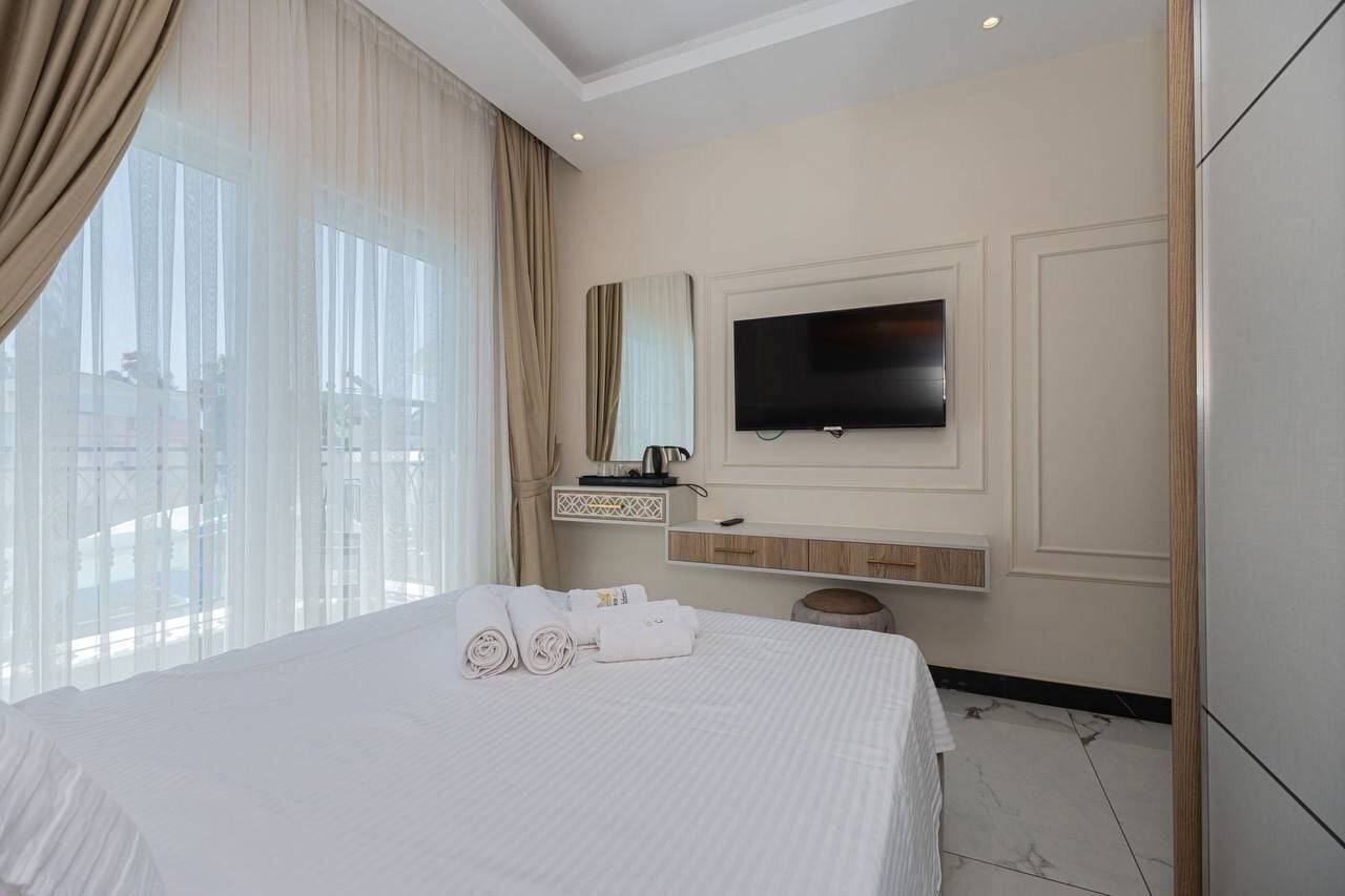 Habitación de hotel con una cama grande, un televisor de pantalla plana en la pared, una mesita con una tetera y vistas a la ciudad a través de amplios ventanales.