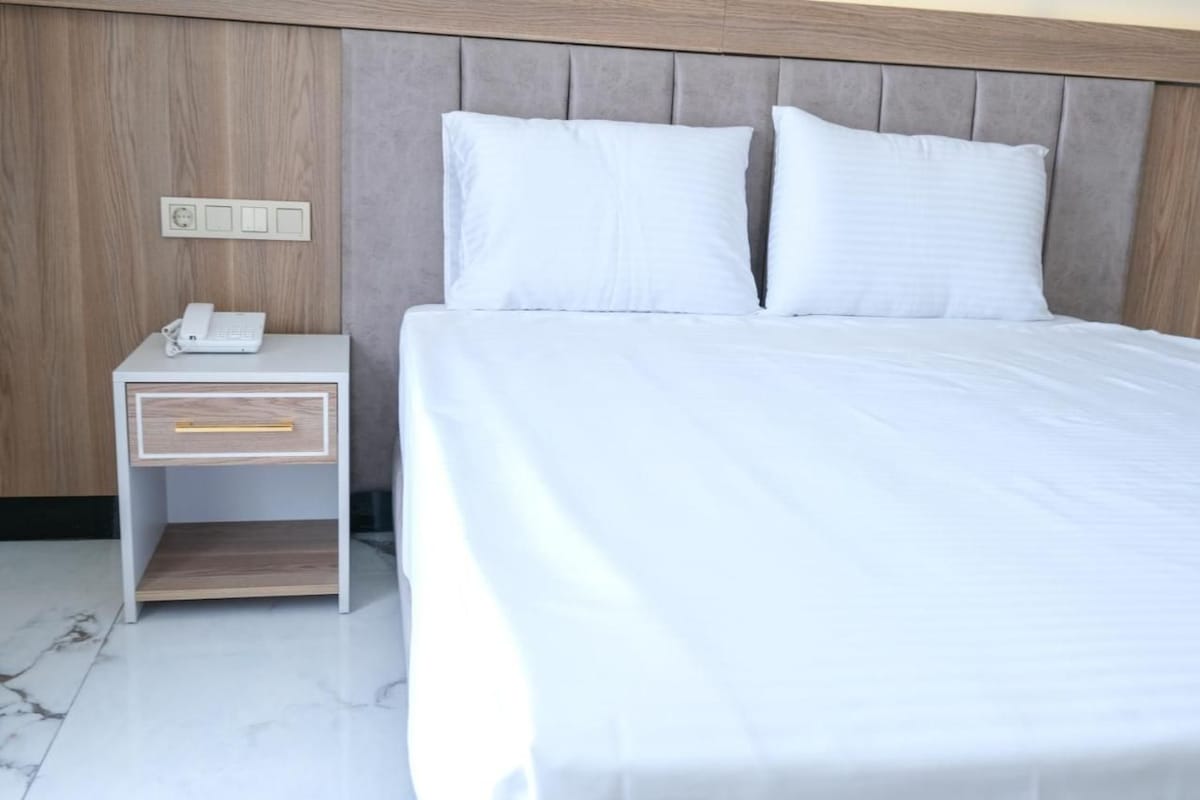 A neatly made bed with white linens, a bedside table with a phone, and a wall-mounted light switch.