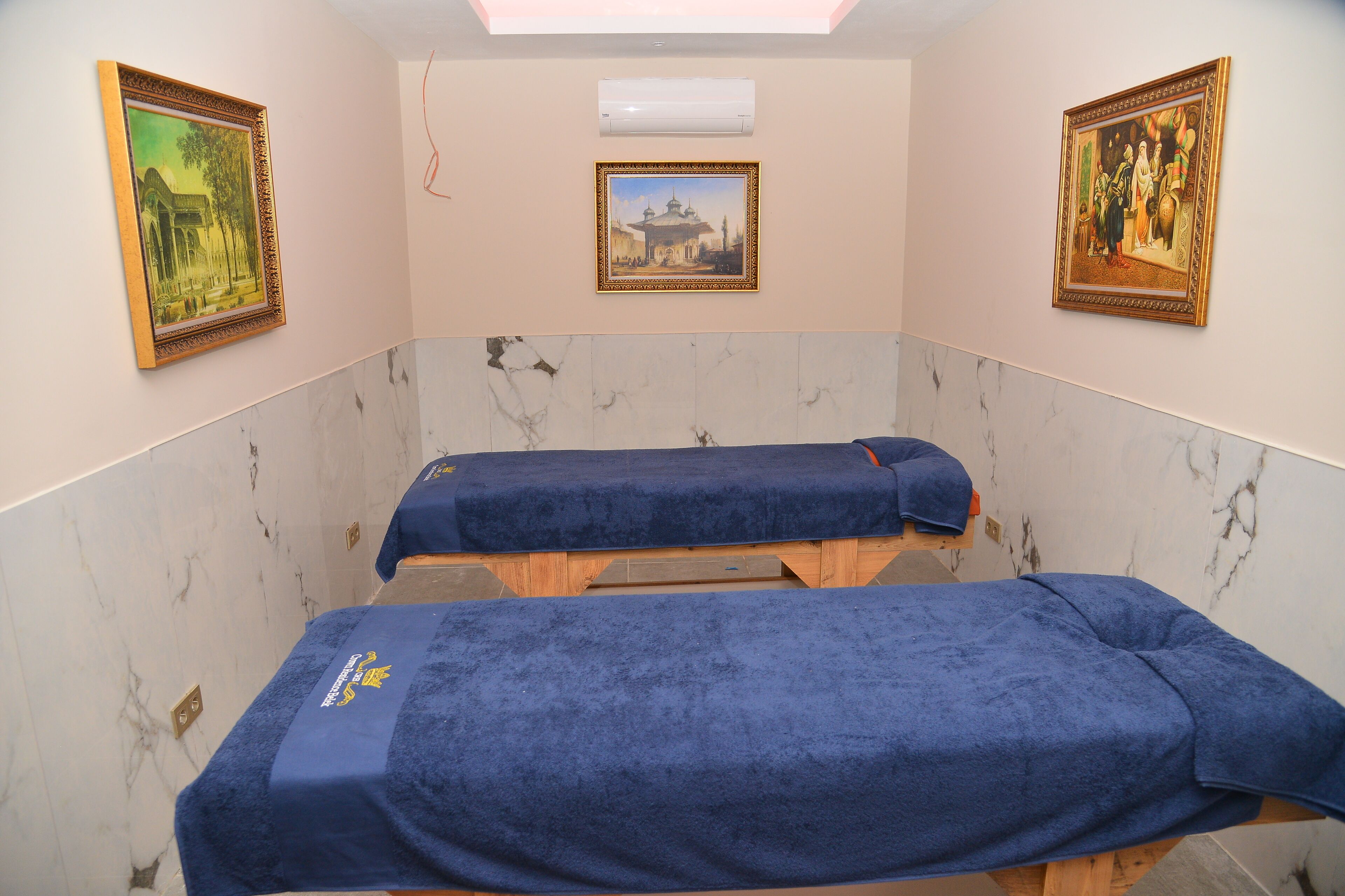 Una habitación con dos mesas de masaje cubiertas con toallas azules, paredes de mármol y dos cuadros enmarcados.