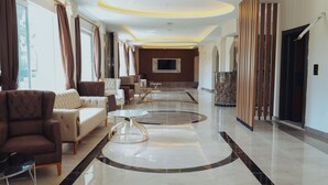Eine geräumige Lobby mit hoher Decke, einer zentralen Sitzecke mit Sofas und Tischen, einem großen Fernsehbildschirm und einem modernen Design mit Holzelementen.