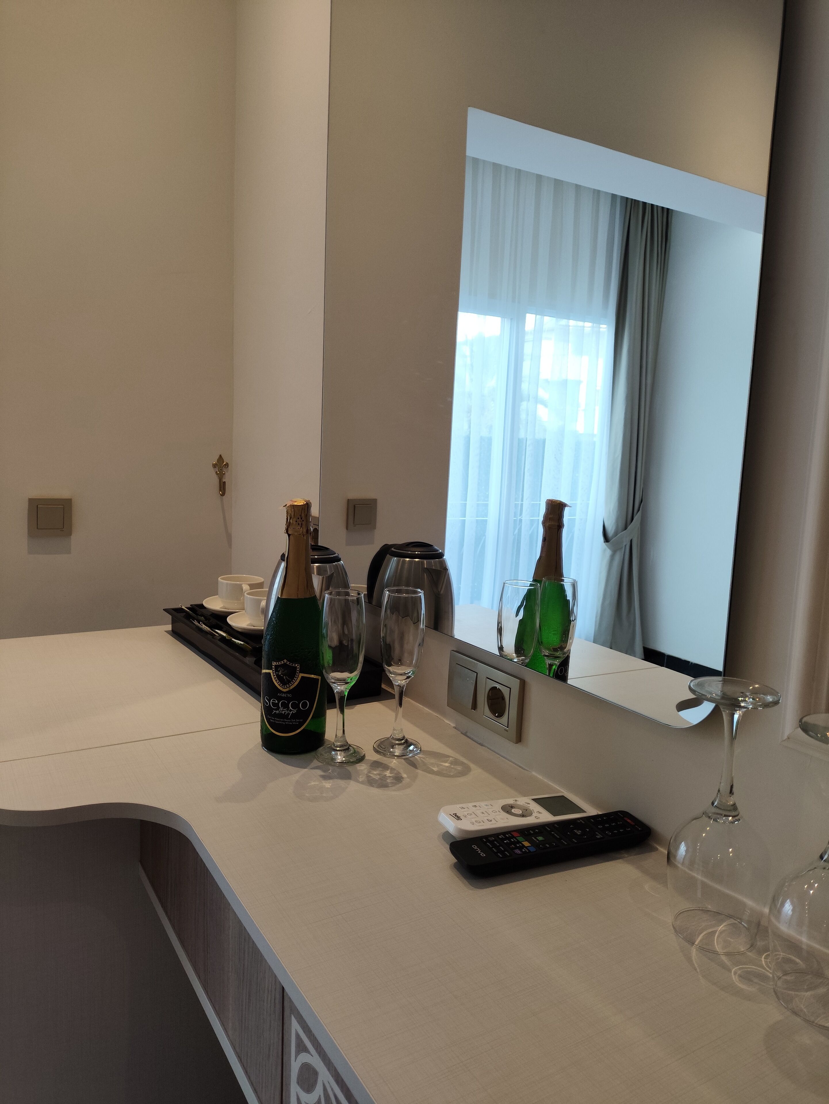 A hotel room with a minibar, a bottle of champagne, two wine glasses, a remote control, and a mirror reflecting the room.