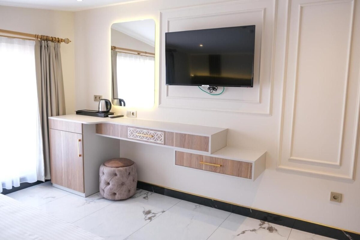 A modern bathroom with a vanity, a mirror, a flat-screen TV, and a round stool.