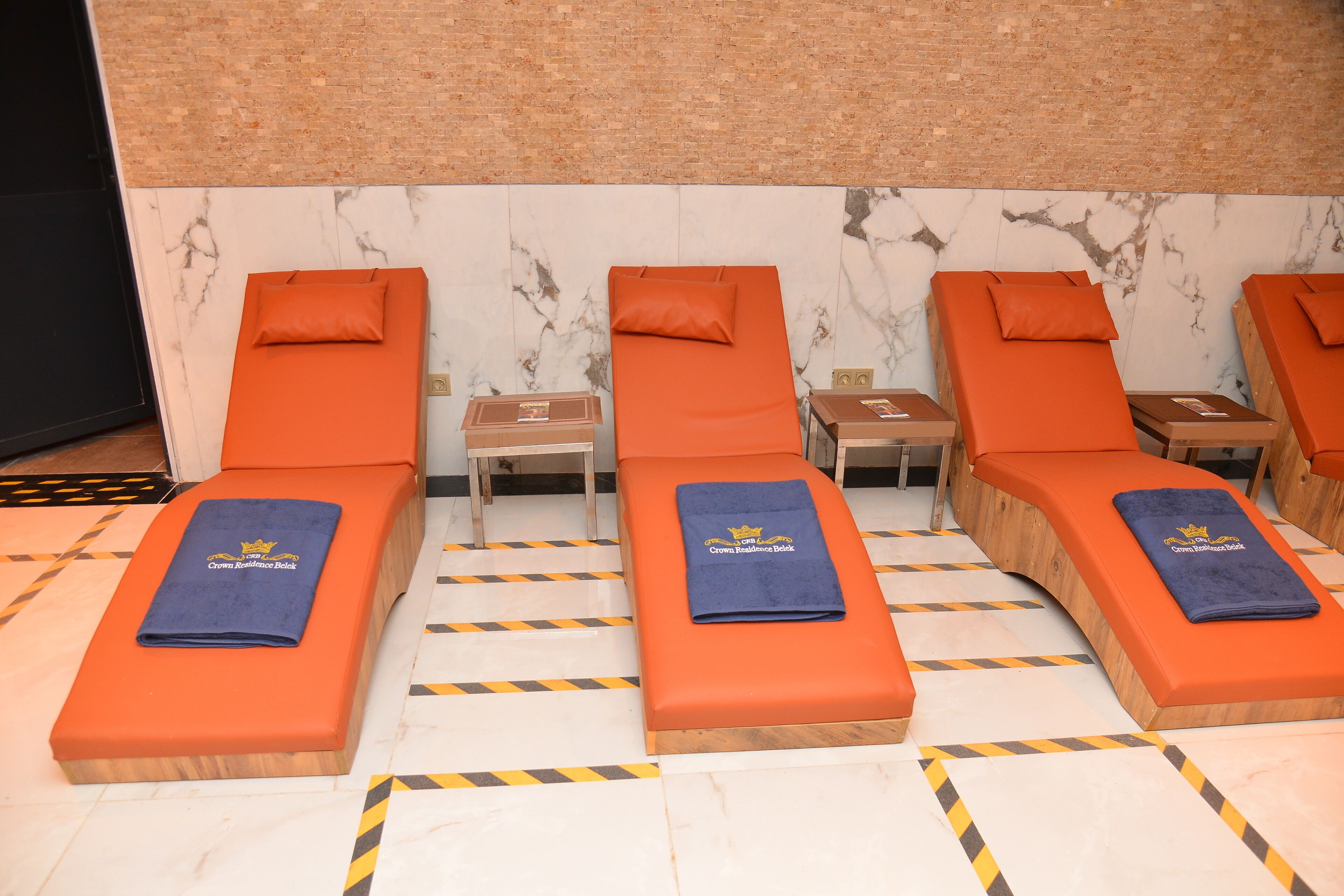 Una fila de tumbonas naranjas con toallas azules, un banco de madera y una pared de mármol con diseño de ladrillo.