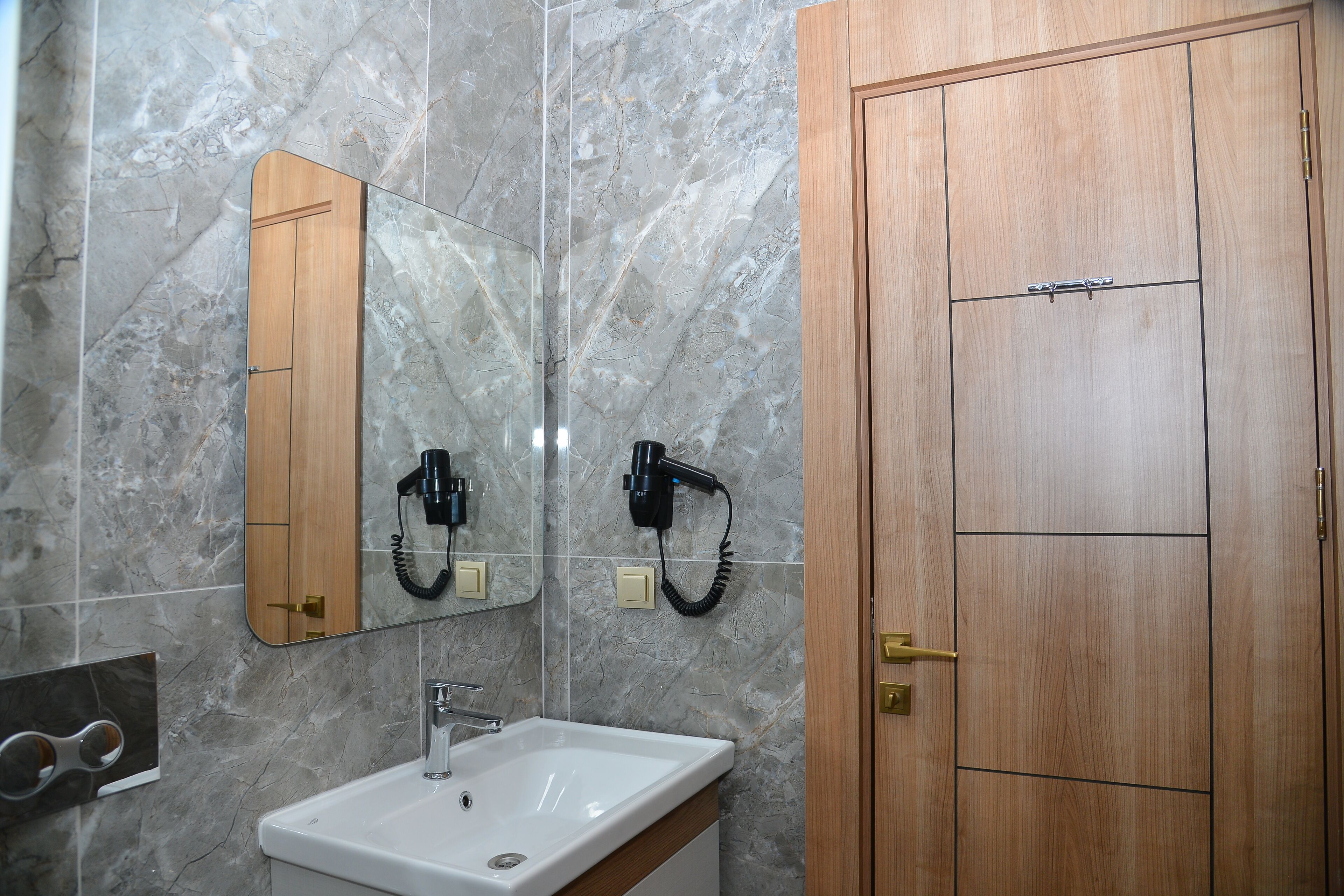 A bathroom with a marble wall, a wooden door, a mirror, a sink, and a hairdryer mounted on the wall.