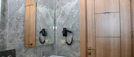 A bathroom with a marble wall, a wooden door, a mirror, a sink, and a hairdryer mounted on the wall.