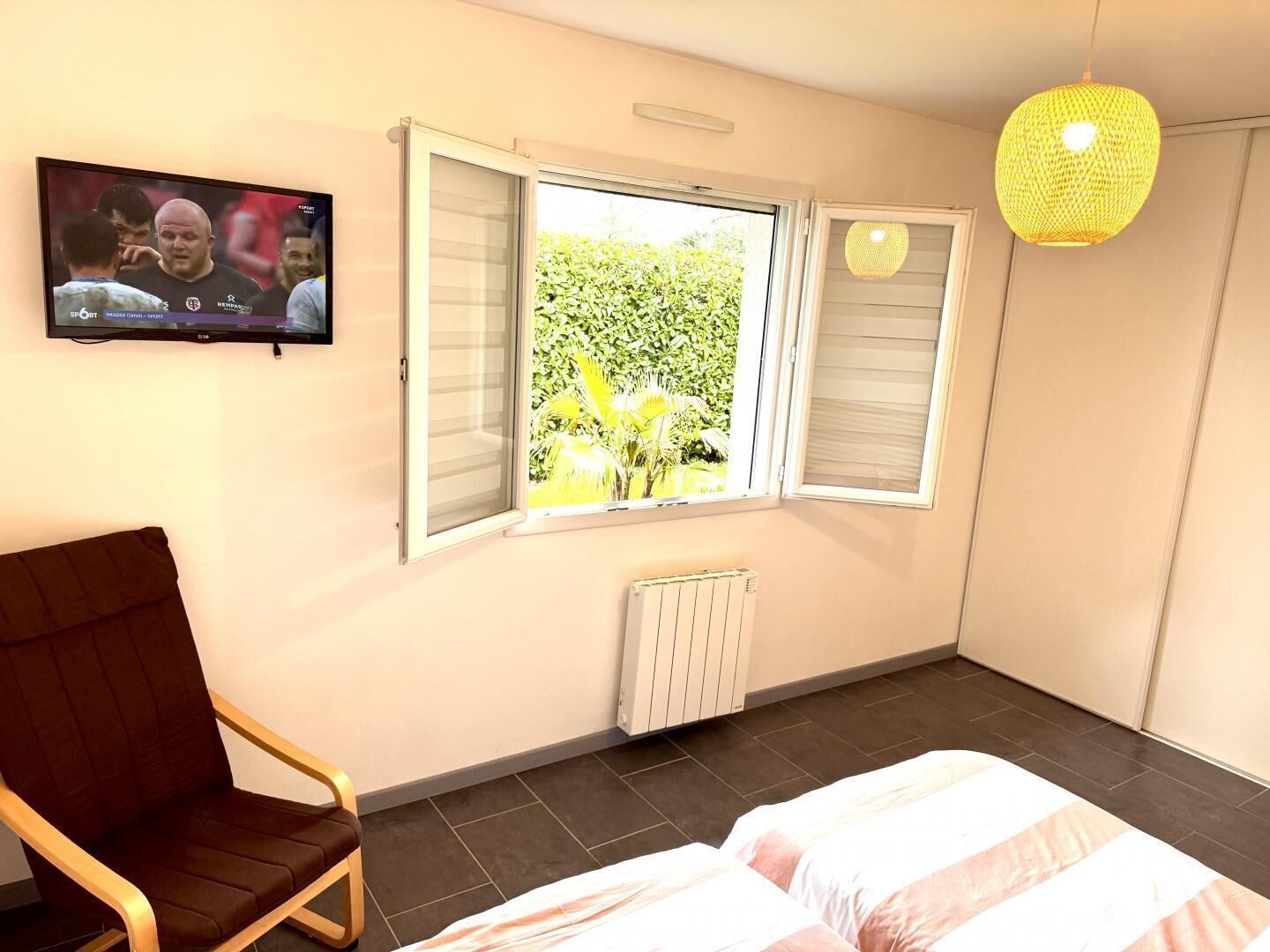 A bedroom with a bed, a chair, a television, and windows with a view of greenery.