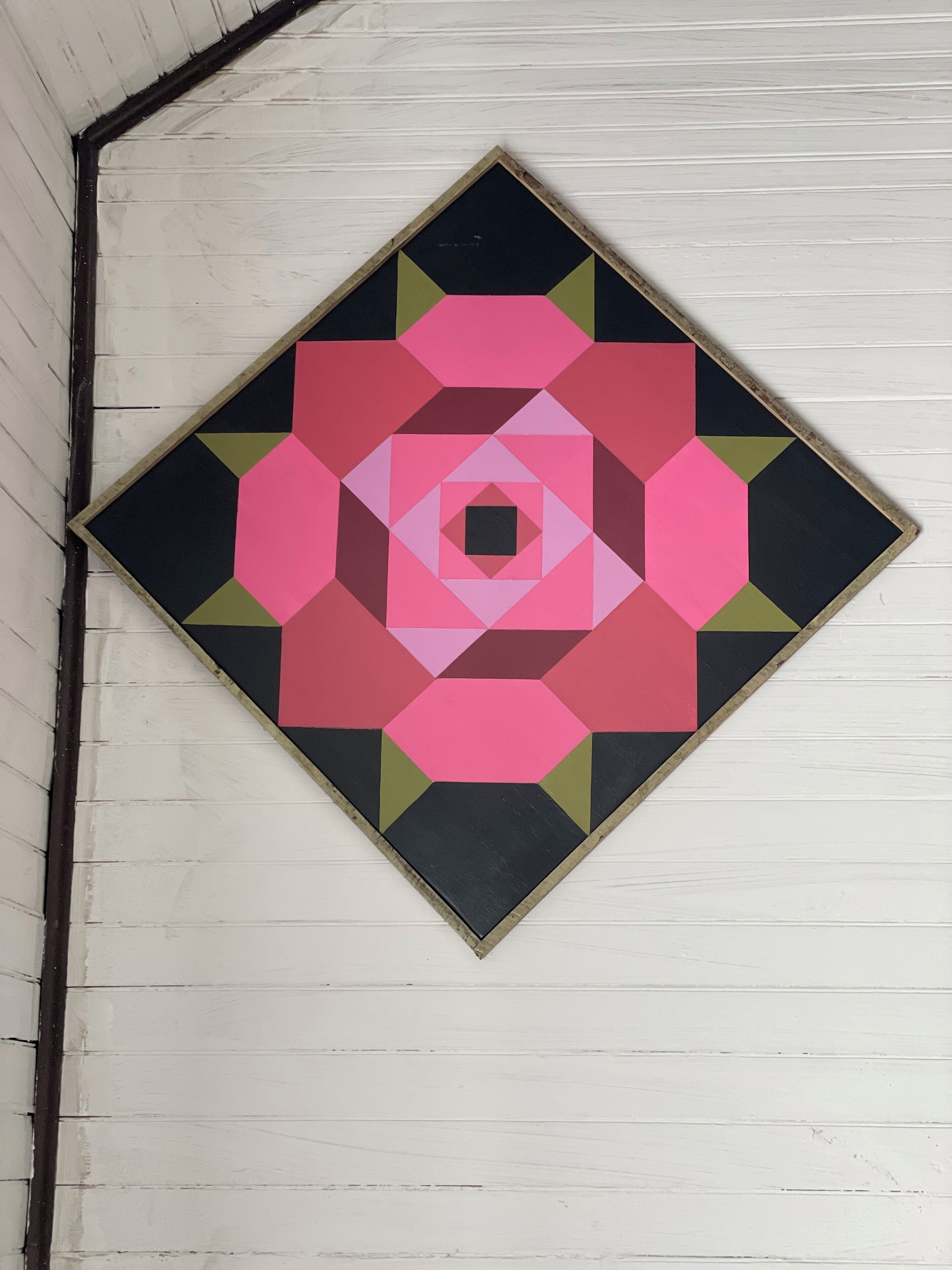 Obra de arte geométrica con forma de diamante, que presenta un hexágono central rodeado de formas concéntricas en rosa, negro y verde.