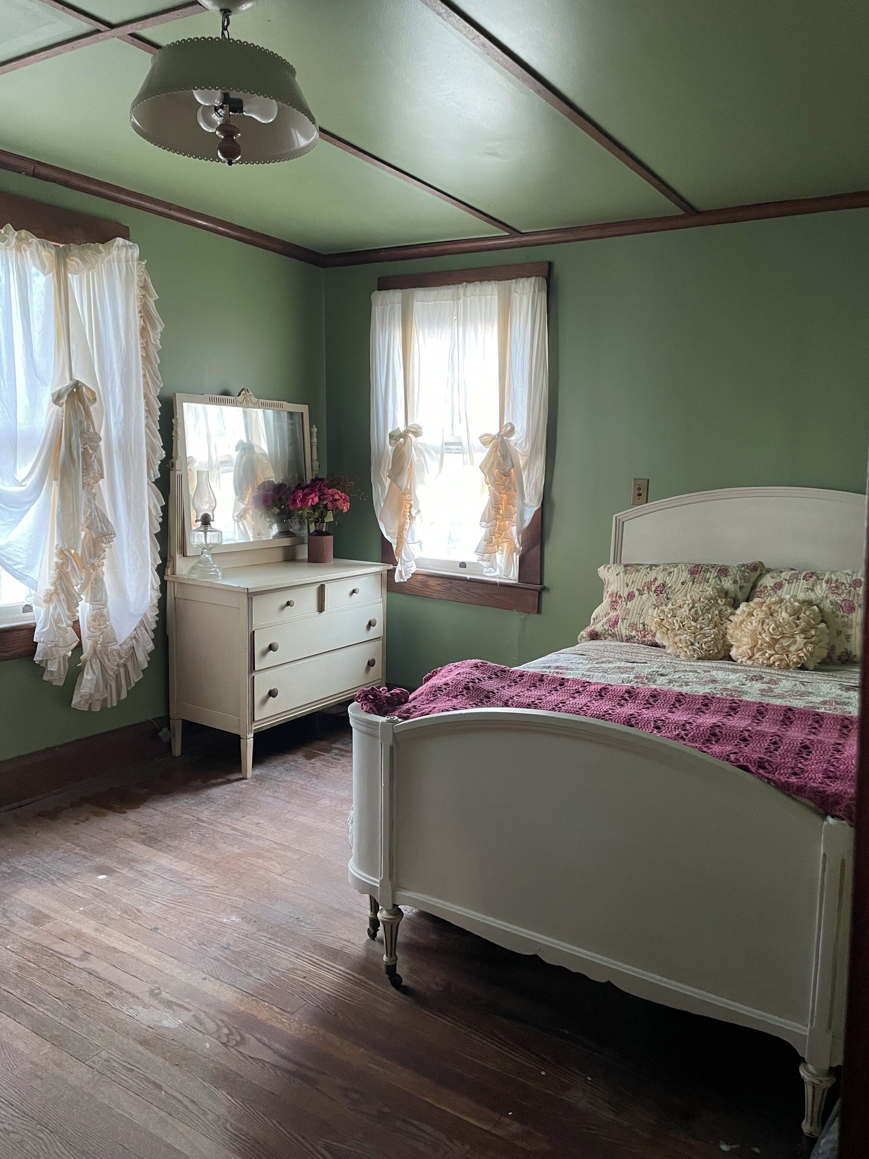 A bedroom with a wooden floor, a white bed with a pink blanket, a dresser with a mirror, and a window with curtains.