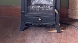 A small, dark-colored wood-burning stove with a glass door, set against a green wall and wooden floor. - Historic Carriage House Suite (Mannington)