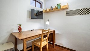 Habitación de esquina con escritorio de madera, silla, televisión, estantería con libros y una planta, y un tablero de ajedrez fijado a la pared.