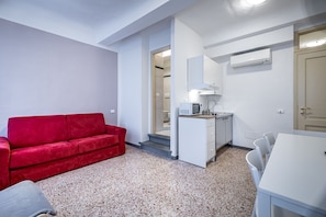 A compact room with a red sofa, a small kitchenette, a white dining table, and a door leading to another room.