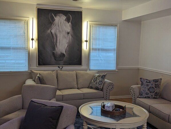 A living room with a large horse painting, two sofas, a coffee table, and a window with blinds.