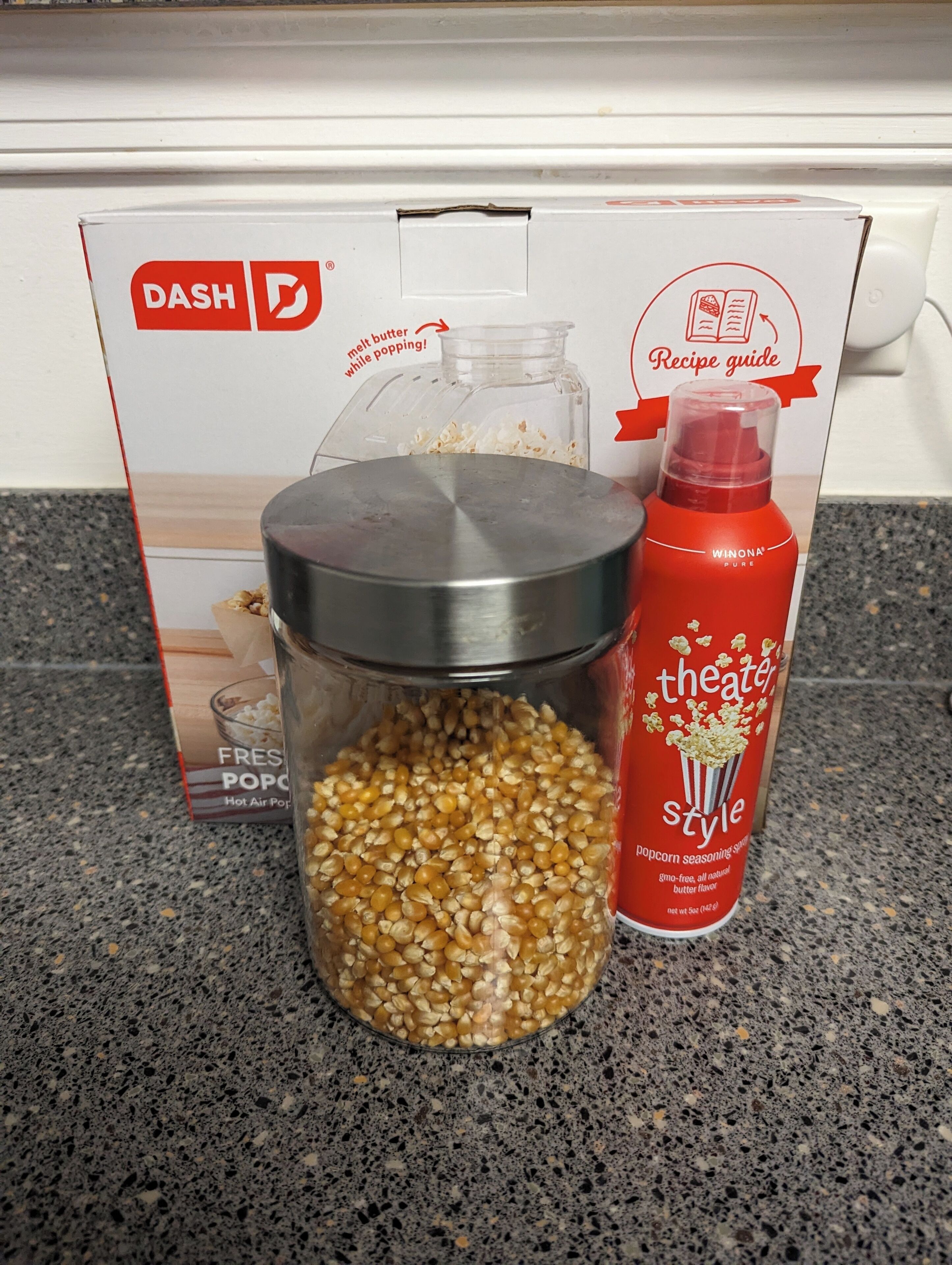 A popcorn maker, a popcorn seasoning bottle, and a box of fresh popcorn on a kitchen counter.