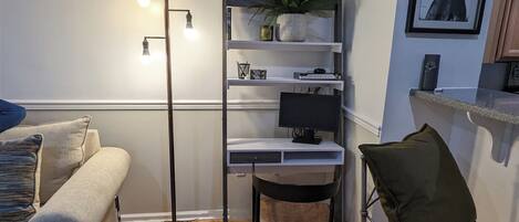 A modern living room with a white sofa, a small round ottoman, a metal-framed desk with a computer, a floor lamp, and a potted plant on a shelving unit.