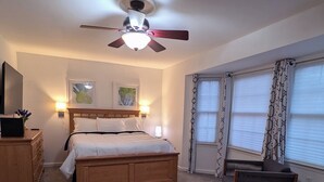 Une chambre à coucher avec un lit, une table de chevet en bois, un bureau avec une lampe, un ventilateur de plafond et une fenêtre avec des rideaux.