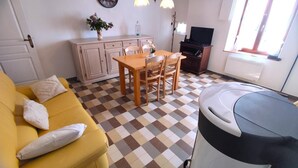 A room with a checkered floor, a yellow sofa with pillows, a wooden dining table with chairs, a cabinet, a television, and a trash bin. - LA MADELEINE - MALANDRY (MALANDRY)