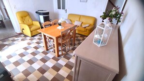 A compact living space with a checkered floor, a yellow sofa, a wooden dining table with chairs, a small table with a vase of flowers, and a white cabinet. - LA MADELEINE - MALANDRY (MALANDRY)
