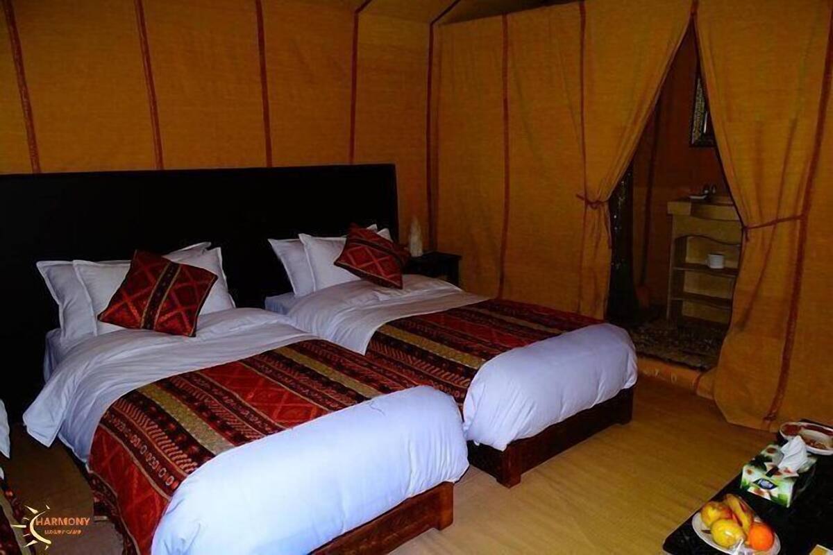 Une chambre avec deux lits, chacun recouvert de draps blancs et d’une courtepointe rouge à motifs. On y trouve une petite table avec des fruits et une bouteille.