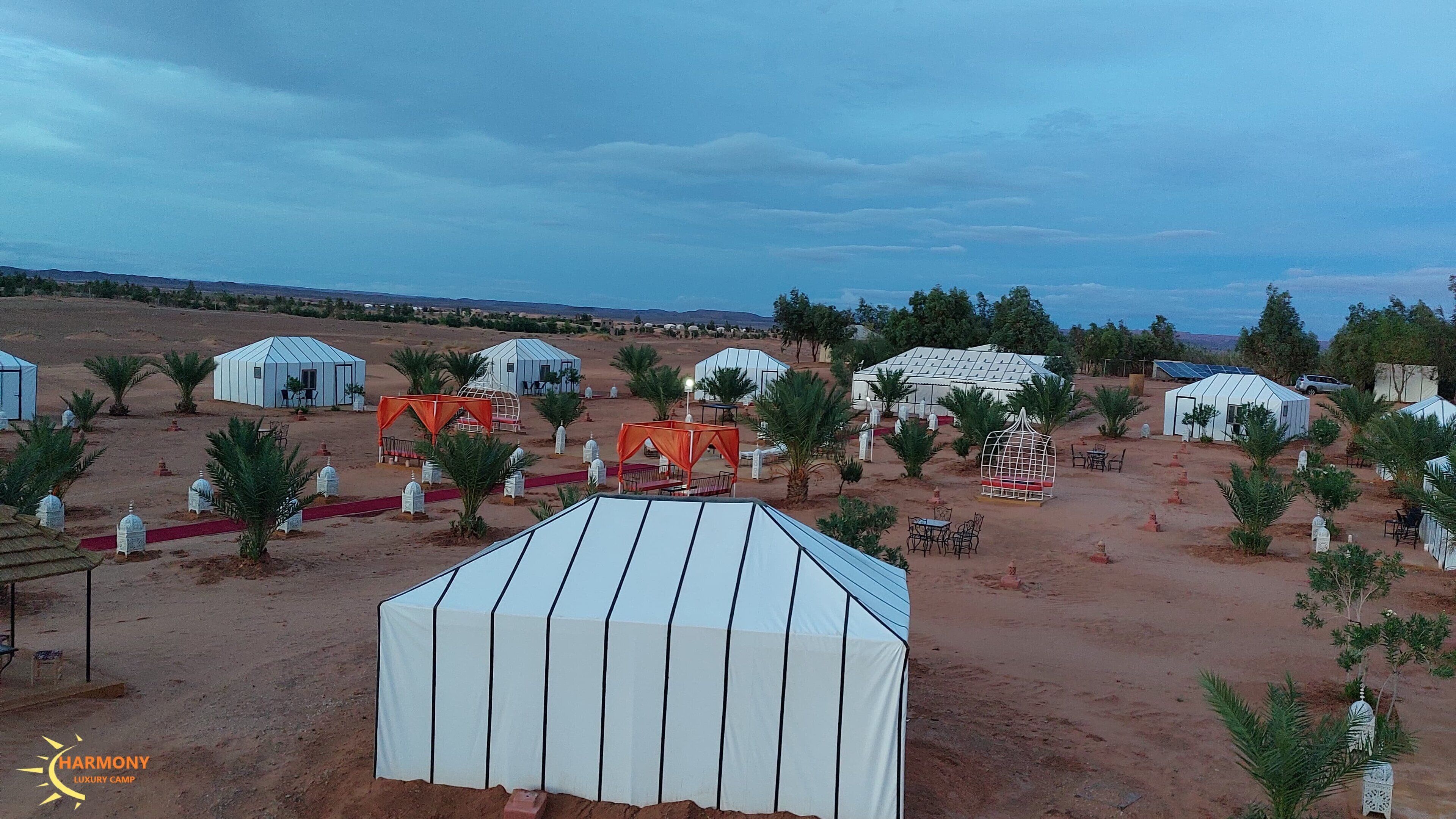 Un campamento en el desierto con varias tiendas de forma abovedada, algunas con toldos rojos, y palmeras.