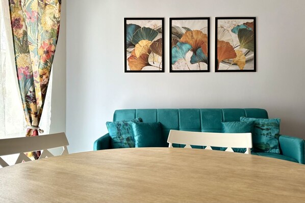 A room with a teal sofa, a wooden dining table, and three framed botanical prints on the wall.