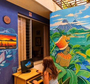 A room with a computer on a desk, a window with blinds, and a mural on the wall depicting a volcano and a person with a headdress. - Hostal los Volcanes ciudad de Guatemala (Guatemala City)