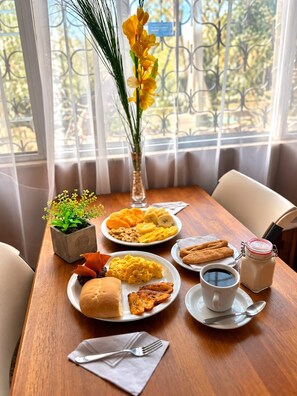 Una mesa de comedor con platos de comida, un florero con flores amarillas y vista a zonas verdes a través de una ventana.