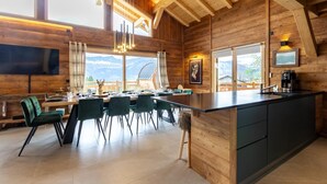 A wooden cabin with a dining area, kitchen, and large windows offering mountain views.