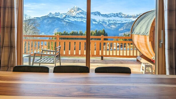 A room with a wooden table and chairs, a wooden deck with a railing, and a view of snow-capped mountains.