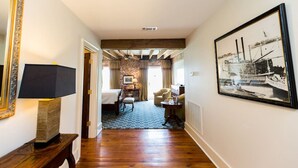 A hotel room with a bed, a desk, a chair, a lamp, a framed picture, and a view of a brick wall and ceiling beams.