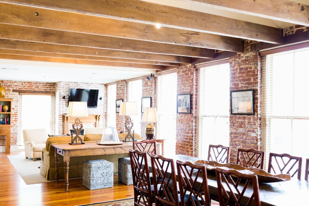 A spacious room with exposed brick walls, wooden beams on the ceiling, a dining table with chairs, a sofa, and a television mounted on the wall.