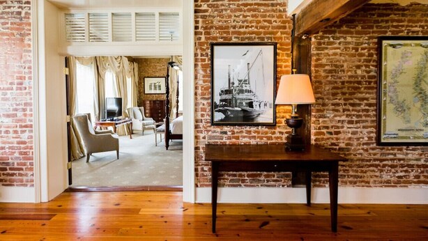 A room with exposed brick walls, a wooden floor, a table with a lamp, and a framed picture on the wall. - The River Walk Inn  - Room 806 (Natchez)