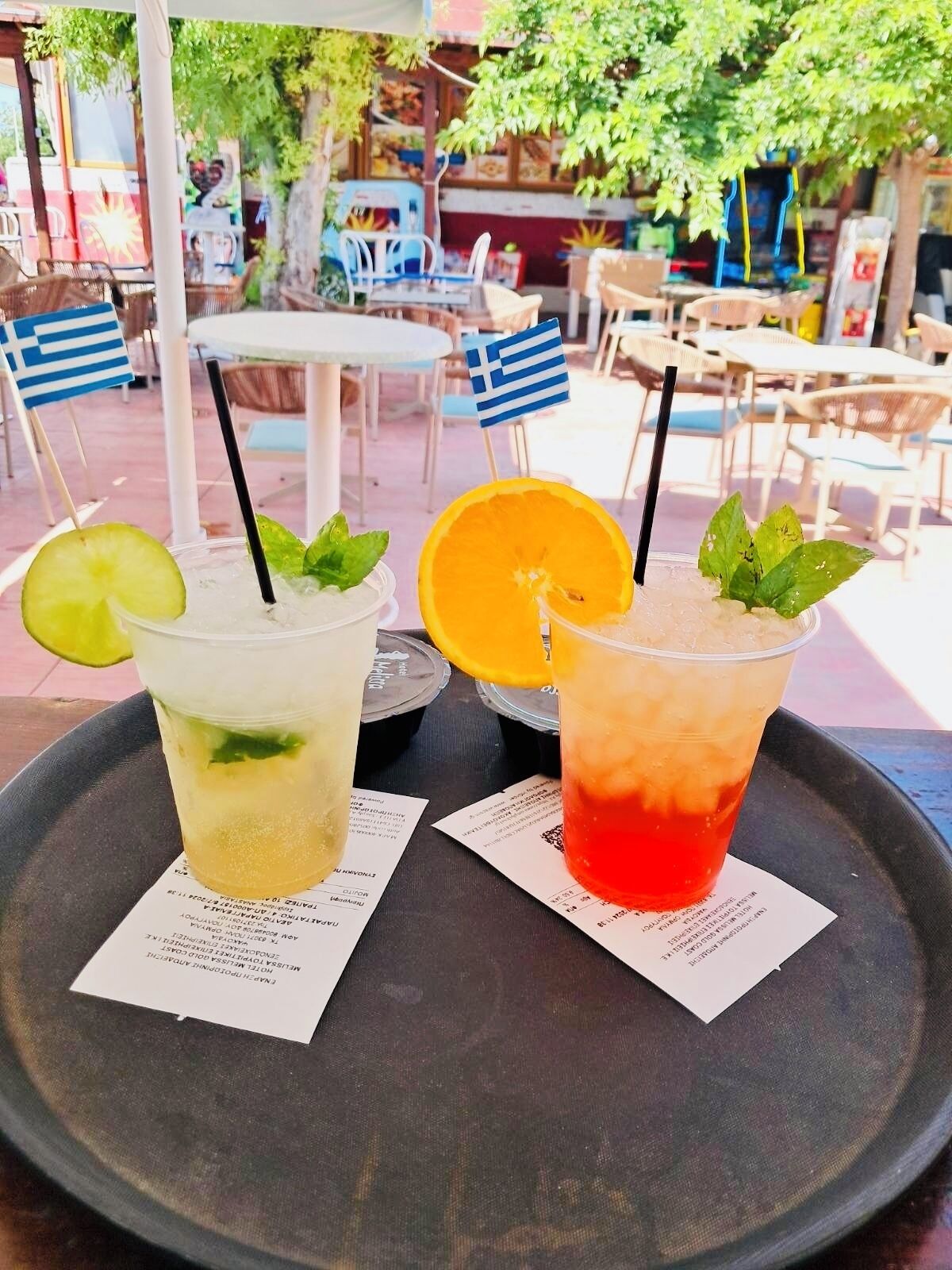 Two drinks on a tray with menus, set in an outdoor seating area with tables and chairs, and a tree in the background.