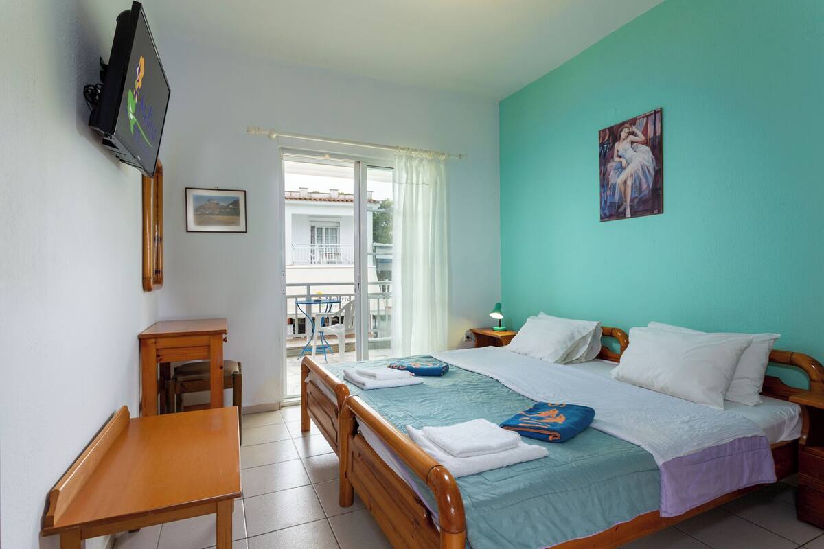 A hotel room with a bed, a desk, a chair, a TV, and a balcony with a view of buildings.