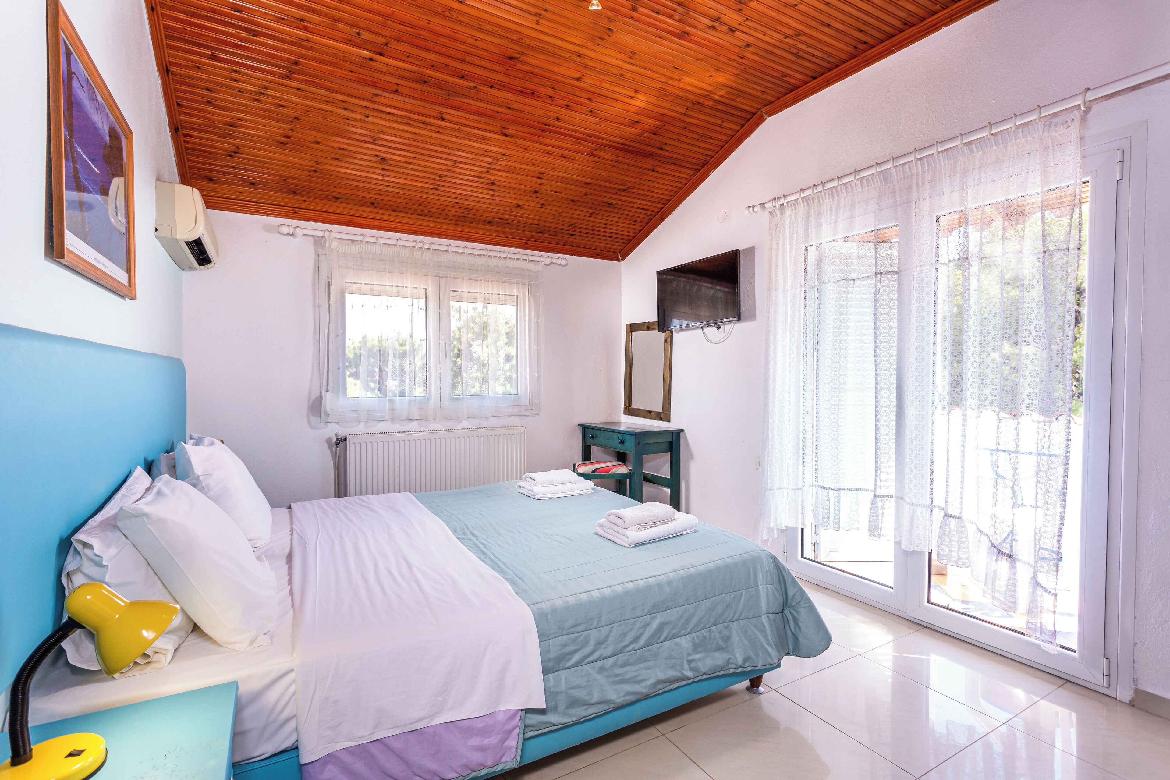 Une chambre avec un plafond en bois, un lit recouvert d’une courtepointe bleue et blanche, une table de chevet avec une lampe, et une fenêtre ornée de rideaux légers.
