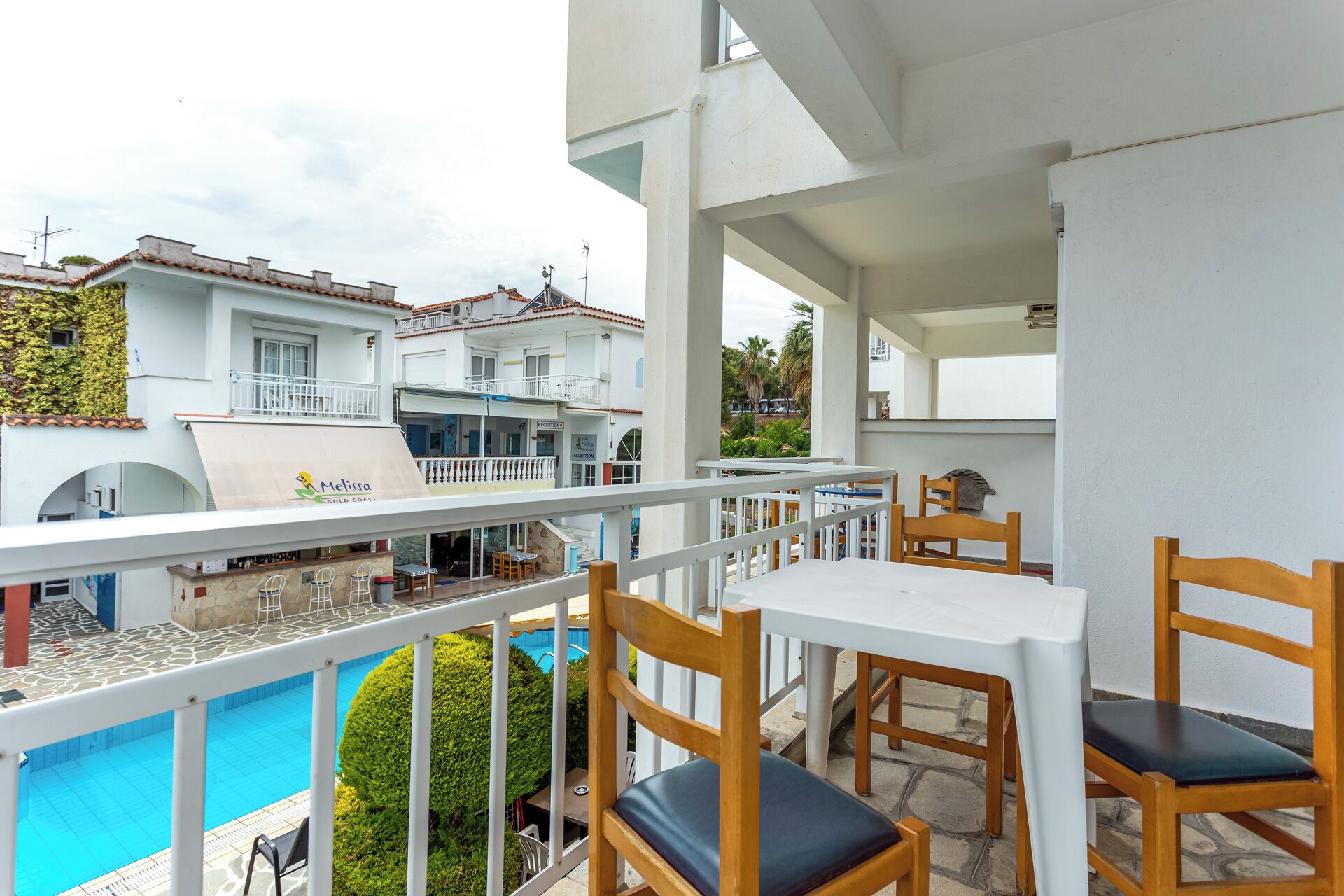 A balcony with wooden chairs and a table overlooking a swimming pool, with buildings in the background.