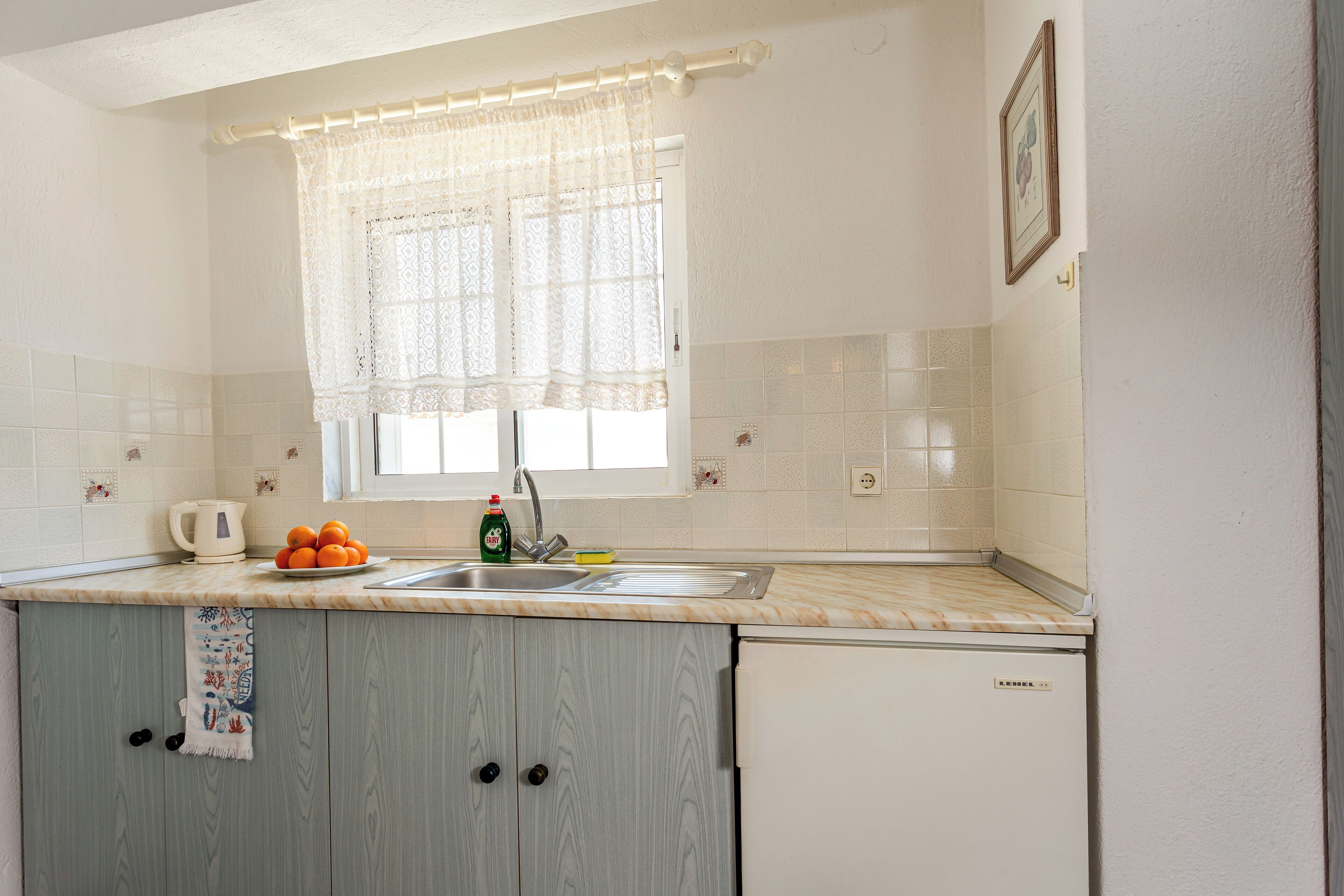 A kitchen with a sink, a window with a curtain, a kettle, and a bowl of oranges.