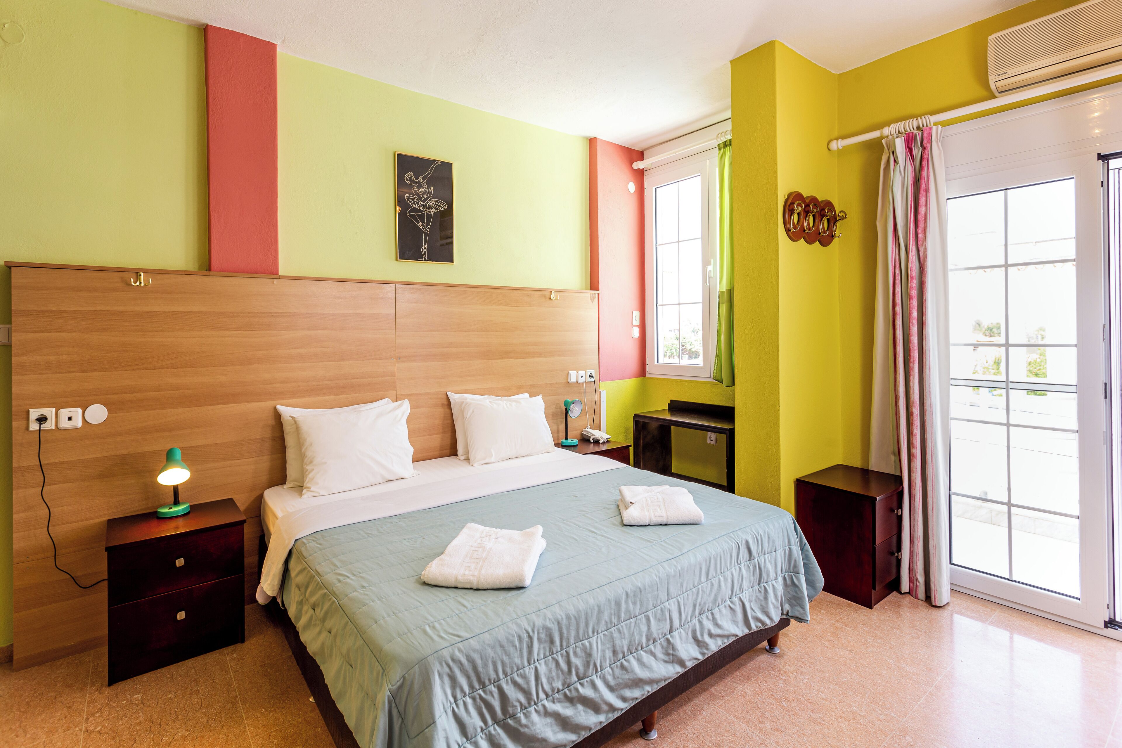Una habitación de hotel con cabecera de madera, cama con ropa de cama blanca, mesita de noche con lámpara y ventana con cortinas.