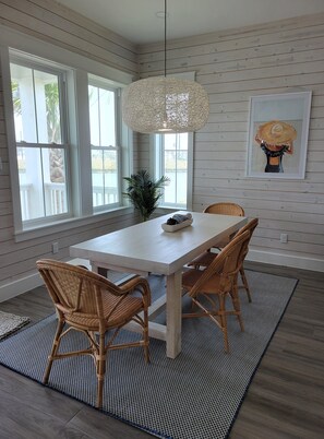 A dining area with a white table and wicker chairs, a hanging light fixture, and a framed artwork on the wall. - The Laguna Lady--Lakefront 3 BR House with Private Dock, Minutes to the Beach! (Port Aransas)