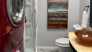 A compact bathroom with a red washing machine, a glass shower enclosure, a white toilet, and a wooden sink with a bowl.