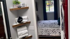 A compact room with a wooden floor, a bed with a patterned bedspread, a red washing machine, a wooden shelf with towels, a mirror, and a small plant.