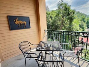 A balcony with a metal table and chairs, a framed moose artwork on the wall, and a view of trees and hills. - Legacy at the Summit - 2 Balconies, Mountain Views, Walk Downtown (Gatlinburg)