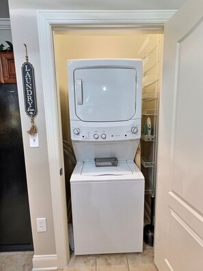 Une petite buanderie comprenant un lave-linge et un sèche-linge, une petite étagère avec des produits de nettoyage et un panneau décoratif sur le thème de la lessive.