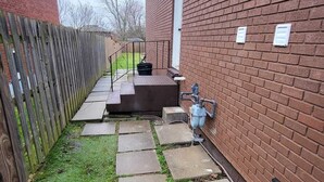 A backyard with a brick wall, a wooden fence, and a concrete pathway leading to a raised platform. - Spacious and Modern Home, 10 mins to Restaurants, Shops and Parks. (Welland)
