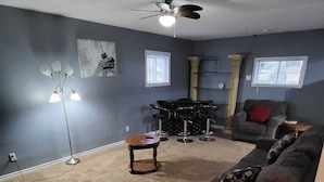 A living room with a dark blue wall, a ceiling fan, a coffee table, a sofa, and a drum set against the wall. - Spacious and Modern Home, 10 mins to Restaurants, Shops and Parks. (Welland)