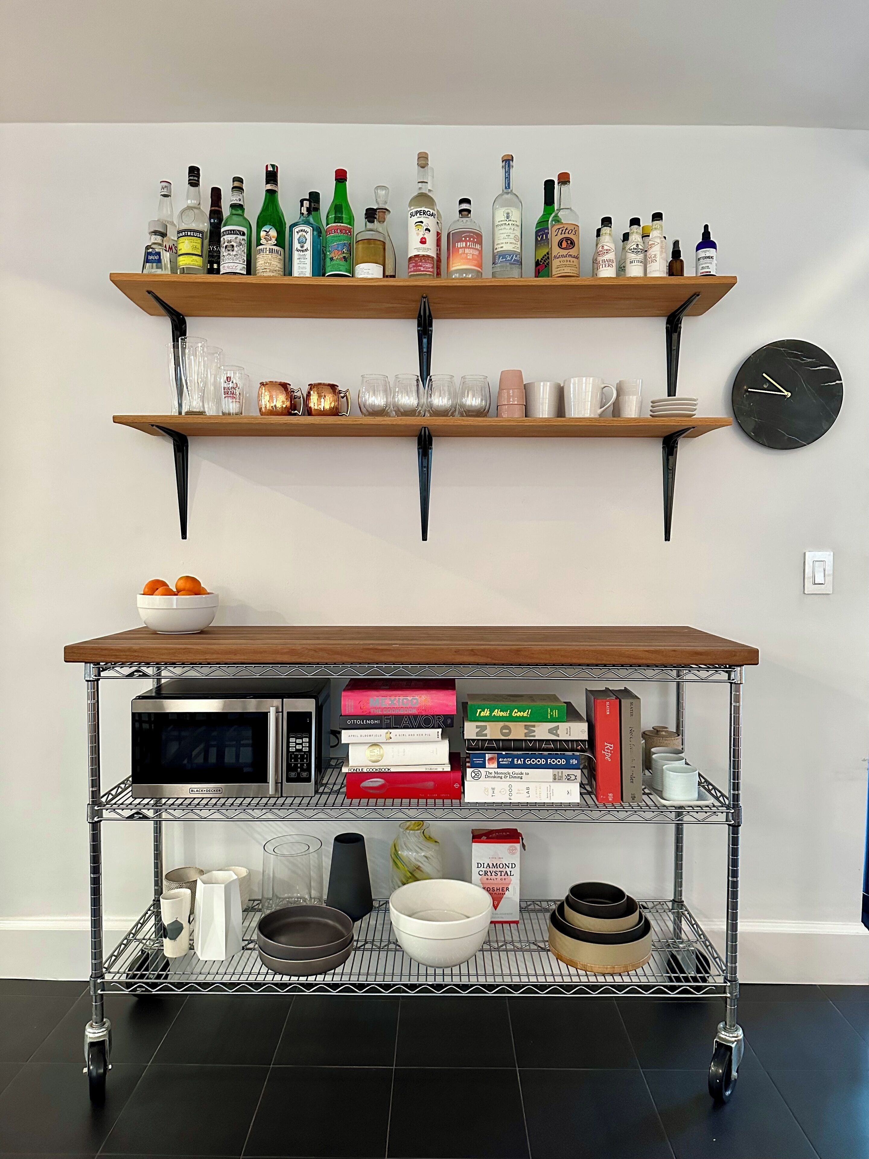 A kitchen with a metal cart holding kitchen appliances and books, a shelf with liquor bottles, and a clock on the wall.